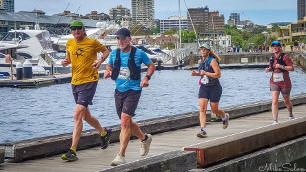 Bondi‑to‑Manly Ultra runners on wooden boardwalk beside Woolloomooloo Bay marina in Sydney, with moored yachts and city buildings in background. [4527]