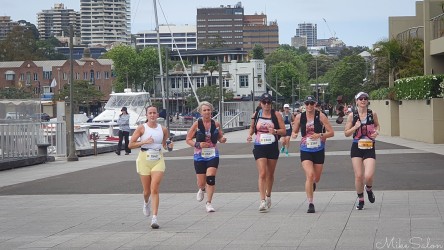 Bondi‑to‑Manly Ultra runners on harbourside path at Woolloomooloo Bay Sydney, with boats and city buildings in background under soft daylight.