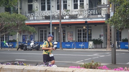 Bondi‑to‑Manly Ultra runner in colourful shirt passing Woolloomooloo Bay Hotel in Sydney, with Bondi‑to‑Manly Walk sign, parked scooters, and flower beds in foreground.