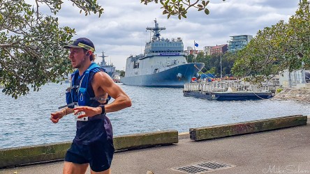 Bondi‑to‑Manly Ultra runner in hydration vest passing Royal Australian Navy ship at Fleet Base East in Woolloomooloo Bay, Sydney, with trees and waterfront in background.