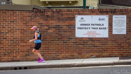 Bondi‑to‑Manly Ultra runner in blue top and pink shoes passing brick wall with Australian Government Department of Defence sign reading "Armed Patrols Protect This Base" at Woolloomooloo, Sydney.