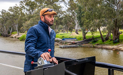 Captain Scott wearing a brown beret and navy jacket, steering the Australian Star paddle steamer along the Murray River near the mooring, surrounded by river gums.