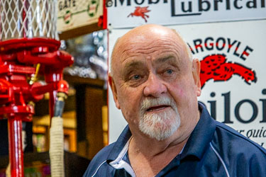 Portrait of Neil, owner of the Great Aussie Beer Shed and Heritage Museum, standing beside vintage Gargoyle Mobiloil signage and red fuel pump.