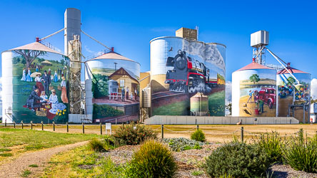 Colbinabbin silo art featuring murals of a picnic scene, railway station, steam locomotive, and vintage farm truck under a bright blue sky.