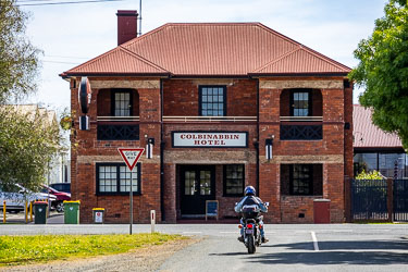 Motorcyclist approaching the two‑storey red‑brick Colbinabbin Hotel with red roof and "Give Way" sign in front, under a clear sky in rural Victoria.