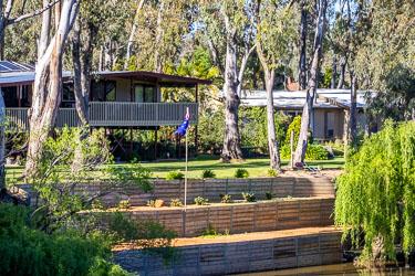 Contemporary riverside house elevated on stilts with a wide deck, surrounded by gum trees and gardens, featuring a solid retaining wall and an Australian flag beside the riverbank.