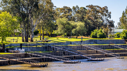 Terraced riverbank with multiple boat ramps and docks leading into the Murray River, bordered by green lawns, large trees, and landscaped gardens behind.
