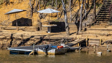 Small aluminium boat moored to a wooden jetty on the Murray River, with two metal umbrellas and a table on a sandy bank backed by timber steps and gum trees.