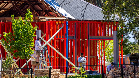 New house under construction with bright red timber framing and corrugated roof, workers on site surrounded by trees and sunlight, symbolising rebuilding after the 2022 Murray River flood.