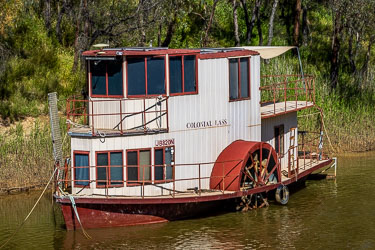 Two‑storey paddleboat named Colonial Lass moored on the Murray River, with white and red exterior, large paddle wheel, and shaded upper deck surrounded by reeds and gum trees.