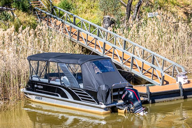 Black pontoon‑style boat named Neptune moored at a floating dock on the Murray River, with a sloped gangway leading up the bank through tall reeds and trees.