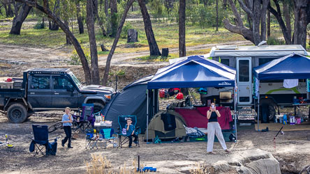 Family campsite beside the Murray River with blue canopy tents, camper trailer, off‑road vehicle, and people relaxing and taking photos among trees and camping chairs.