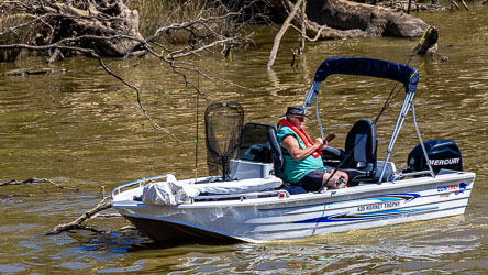Man sitting in a small motorboat named 435 Hornet Trophy on the Murray River, wearing a red life jacket and hat, looking at his phone beside fishing gear and calm brown water.