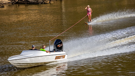 White speedboat named Sinister towing a water‑skier in pink gear on the Murray River, creating a wake across calm brown water with trees along the bank.