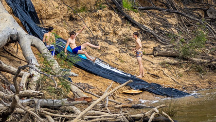 Three boys enjoying a homemade water slide made from black plastic on a sandy Murray River bank, surrounded by tree roots and branches.
