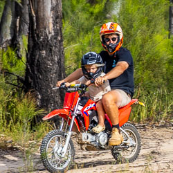 Man and child riding a small red dirt bike on a bush trail, both wearing helmets, surrounded by trees and green vegetation.