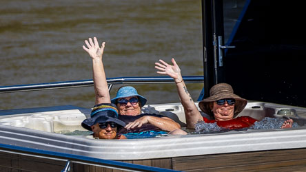 Three mature‑aged people relaxing in a hot tub on a houseboat, wearing sun hats and sunglasses, smiling and waving toward the camera with the Murray River in the background.