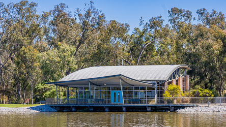 Modern curved‑roof building of the Torrumbarry Weir Interpretive Centre surrounded by river gums, viewed from across the Murray River under clear blue sky.