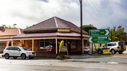 Small corner cafe with red roof, cars parked outside, and road signs pointing toward nearby towns.