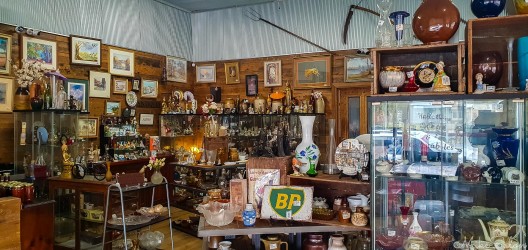 Interior of shop with wooden walls, glass display cabinets, and assorted vintage items including ceramics and framed pictures.