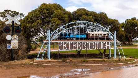 Large metal Peterborough sign with model train and railway crossing signals beside trees.