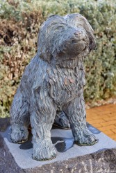 Statue of dog sitting upright on pedestal with greenery in background.