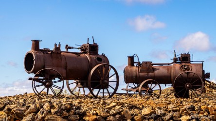 Large rusted cylindrical boiler with wheels on rocky ground under blue sky.