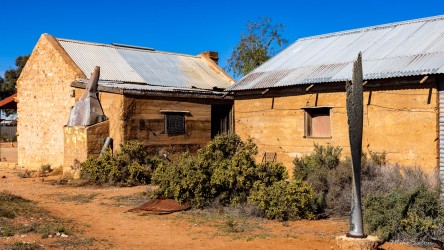 Stone building with metal roof and upright propeller in front, surrounded by scrub and sand.