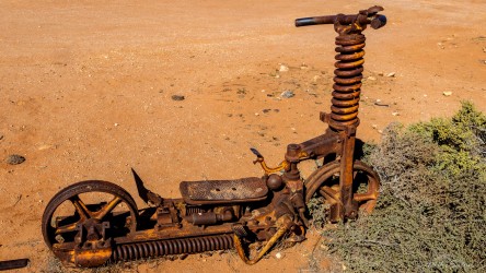 Rusted scooter frame lying on sandy ground with spring suspension and corroded metal parts.