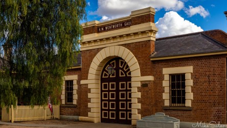 Historic brick jail building with arched doorway, cream stone detailing, and trees beside fence under partly cloudy sky.