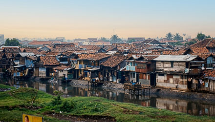 Riverside settlement with wooden houses and tiled roofs reflected in calm canal water under soft morning light in Jakarta.