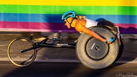 Female wheelchair racer in white top and blue helmet pushing a black Honda racing chair past a rainbow‑striped barrier at the Sydney Marathon 2025, captured in warm morning light.