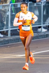 Elite marathon runner Mesfin in orange and white NN Running Team kit, captured mid‑stride on the Sydney Marathon 2025 course near Taylor Square, with crowd barriers and spectators in the background.