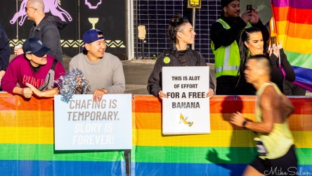 Spectators behind a rainbow‑striped barrier at the Sydney Marathon 2025 holding signs reading "Chafing is temporary, glory is forever" and "This is a lot of effort for a free banana," cheering runners near Taylor Square.