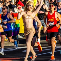 Drag queen in gold outfit and heels holding a red megaphone cheering marathon runners at Taylor Square during the Sydney Marathon 2025, with rainbow banner and spectators in background.