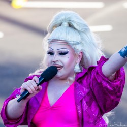 Drag queen MC with platinum blonde hair and sparkling pink outfit holding a microphone and raising an arm while hosting Sydney Marathon 2025 festivities at Taylor Square.