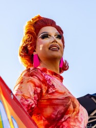 Drag queen with bright red hair and pink‑patterned outfit posing confidently in sunlight at Taylor Square during the Sydney Marathon 2025, with blue sky background.