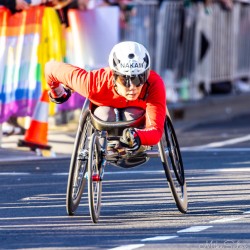 Wheelchair racer Nakam in red top and white helmet pushing hard along Sydney Marathon 2025 course near Taylor Square, with rainbow barrier and spectators in background.