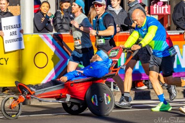 Runner in bright multicoloured shirt pushing a red adaptive racing cart with participant in blue hoodie along Sydney Marathon 2025 course near Taylor Square, with crowd and rainbow banner in background.