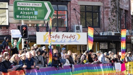 Crowd of spectators behind rainbow banners at Taylor Square during the Sydney Marathon 2025, with Flinders Street road sign and Cafe Freda