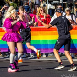 Drag queen in bright pink outfit cheering and high‑fiving marathon runners at Taylor Square during the Sydney Marathon 2025, with rainbow banner and spectators in background.