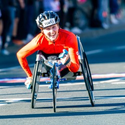 Wheelchair racer Willi in orange racing kit and black helmet smiling while pushing hard along Sydney Marathon 2025 course near Taylor Square, with crowd in background.