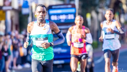 Elite marathon runner Gobena in white and turquoise kit running through Taylor Square during the Sydney Marathon 2025, with timing display and other runners in background.