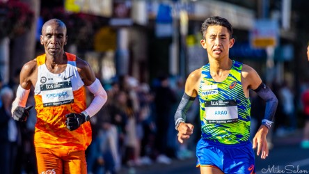 Elite marathon runners Eliud Kipchoge in orange and Masato Arao in blue‑green kit running side by side through Taylor Square during the Sydney Marathon 2025, with crowd in background.