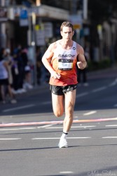 Australian marathon runner Fraser Darcy in white and orange RunAsOne kit running through Taylor Square during the Sydney Marathon 2025, focused and determined under morning light.