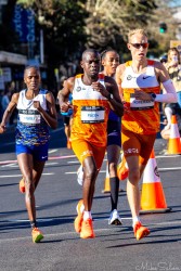 Elite marathon runner Brigid Kosgei in blue and white kit running with two pacers in orange INEOS uniforms through Taylor Square during the Sydney Marathon 2025, with spectators and traffic cones in background.