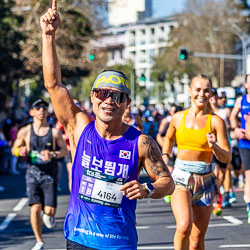 A Korean runner bursts into a smile as he passes the Taylor Square cheer squad, raising a finger skyward in celebration. His energy and colour embody the marathon’s inclusive, global spirit. [3993]