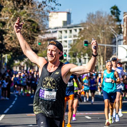 A runner possibly from the Bronx Burners team lifts his arms in delight as he passes through Taylor Square, responding to the cheers and colour of the crowd. The moment radiates shared joy and the universal thrill of endurance. [3996]