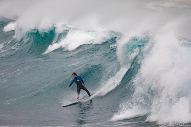 A surfer in a dark wetsuit riding down the face of a large, stormy wave as white spray crests high above him at Bronte Beach.