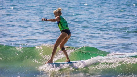 A female surfer in a green rash vest balancing near the front nose of her longboard as she rides a gentle green ocean wave.